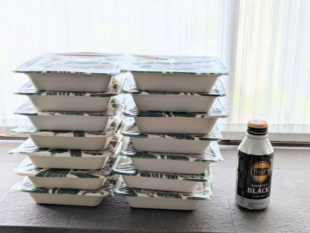 三ツ星ファームの冷凍弁当14食分を積み重ねて撮影。タリーズの缶コーヒーと並べ、容器の大きさや冷凍庫収納時の目安が分かるようにした比較画像。