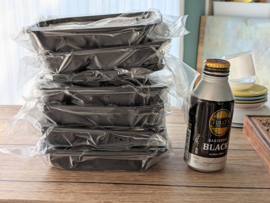 筋肉食堂DELIの冷凍弁当が真空パック状態で7個積まれている。隣にTULLY'Sの缶コーヒーが置かれてサイズ比較になっている。