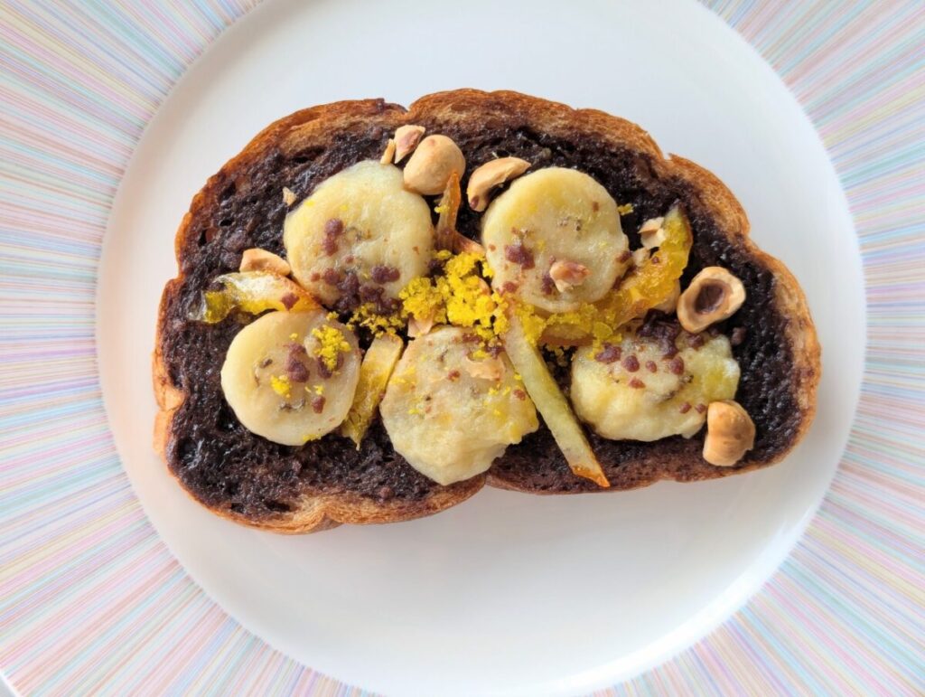 トーストしたパンにビターなチョコクリームを塗り、バナナ・オレンジピール・ナッツ・黄色のパウダーをトッピングしたオープンサンド。オレンジリキュールの香りがアクセント。白い皿に盛り付けられている。