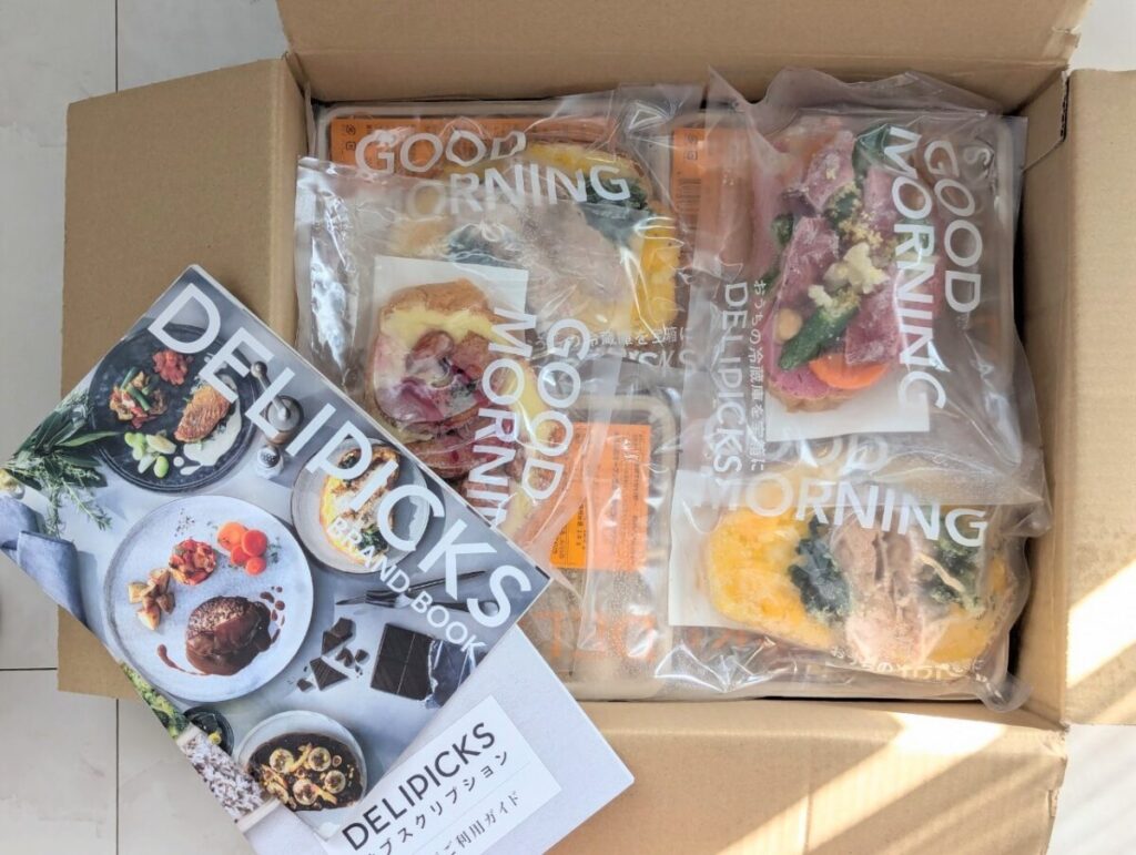 DELIPICKSのオープンサンド4種類とブランドブックが段ボールに入って届いた様子。パッケージには「GOOD MORNING」の文字がプリントされている。