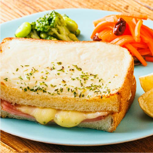 三ッ星ファームの冷凍惣菜「クロックムッシュ」。ハムとチーズをサンドしたトーストの上にホワイトソースとチーズをのせ、パセリをトッピング。付け合わせにブロッコリーと枝豆のソテー、キャロットラペ、ポテトが添えられている。