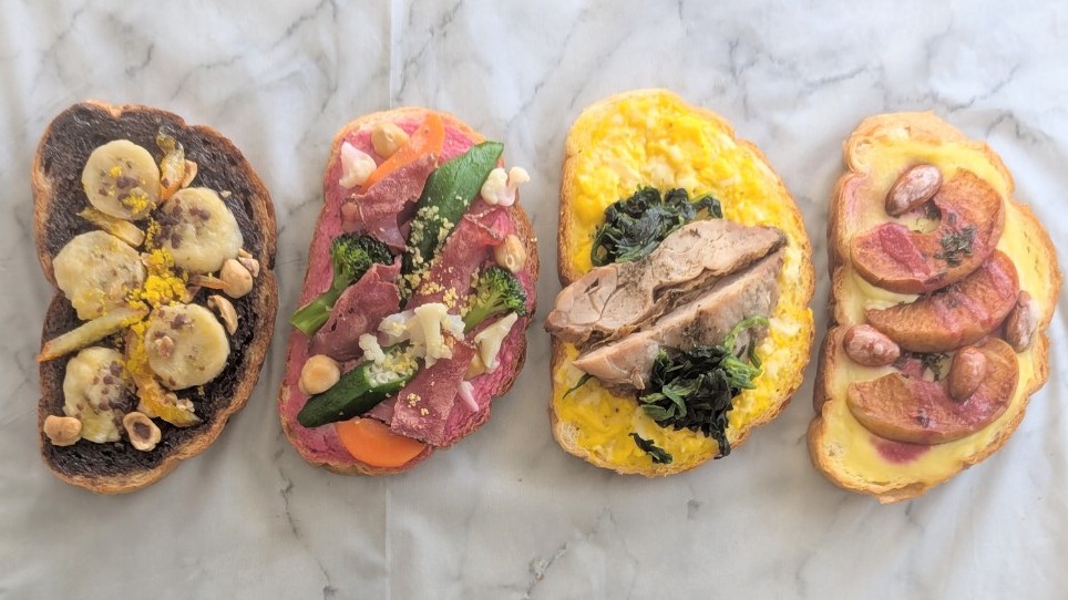 白い大理石の上に並べられたDELIPICKSのオープンサンド全4種。左から順に、バナナとチョコのスイーツ系、彩り野菜とパストラミのサラダ系、ほうれん草とハーブチキンの卵トースト、リンゴのトースト。具材がたっぷりのっていて、見た目も華やか。