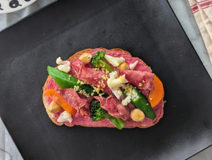 トーストしたパンの上に、クミンが香るビーツフムスを塗り、パストラミビーフと彩り豊かな野菜（ズッキーニ、ブロッコリー、にんじん、カリフラワー、ひよこ豆など）をトッピングしたオープンサンド。黒いプレートに盛り付けられている。