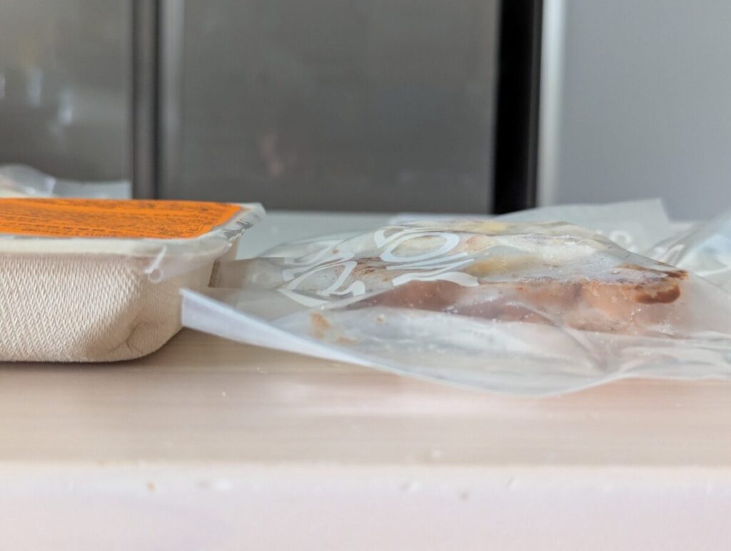 テーブルの上に置かれたDELIPICKSの冷凍オープンサンドと紙製容器のパッケージを横から撮影。冷凍状態の質感や厚みがわかるクローズアップ。