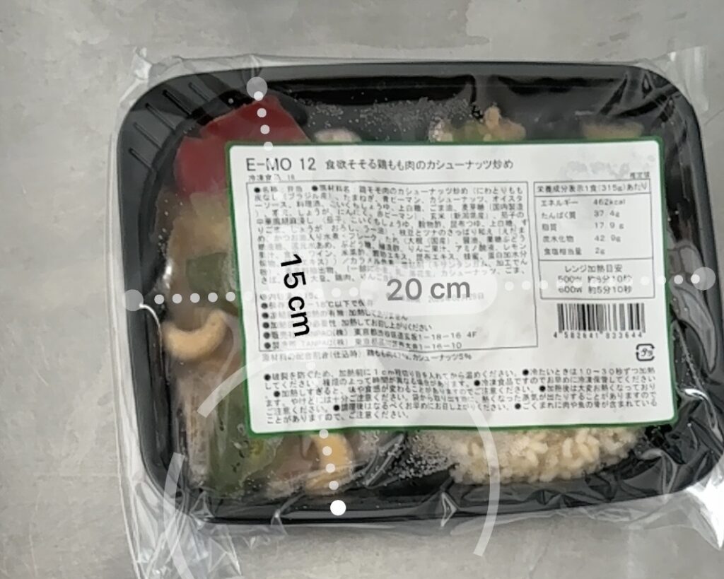 筋肉食堂DELIの冷凍弁当容器を定規で測った写真。黒いトレーの上に透明フィルムがかけられ、ラベルには「鶏もも肉のカシューナッツ炒め」と記載。サイズは横20cm、縦15cmで、一般的な冷凍庫にも収まりやすい大きさ。