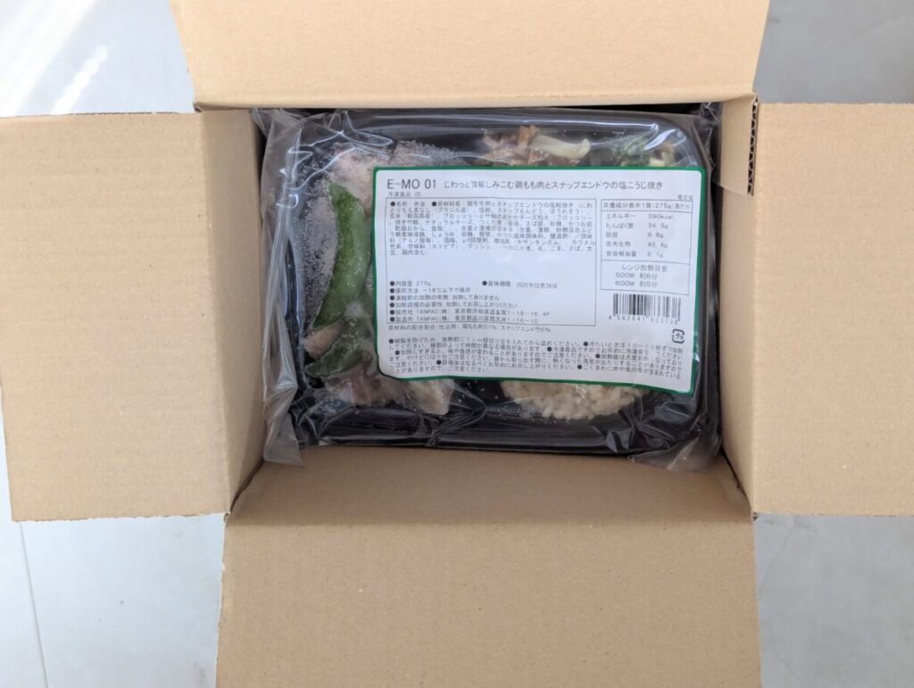 筋肉食堂DELIの段ボールを開けた様子。中にはラベル付きの冷凍弁当トレーが入っており、原材料名や栄養成分、調理方法などが記載されたシールが貼られている。