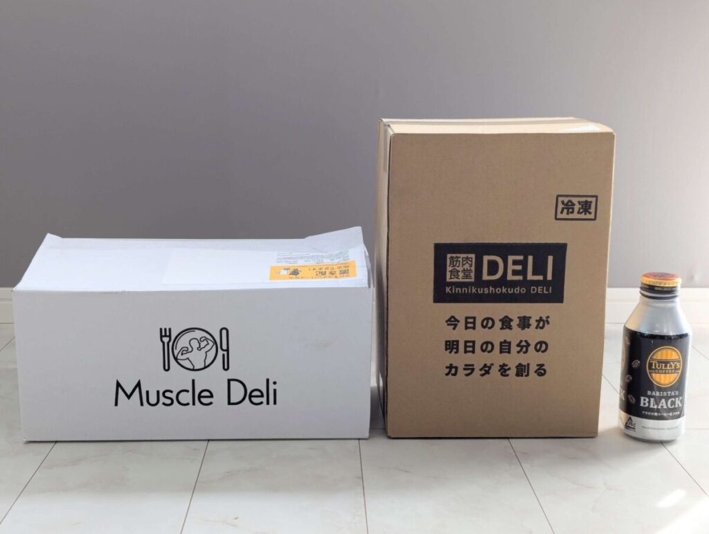 宅配弁当サービスの配送ボックス比較写真。左に白い「Muscle Deli」の箱、右に茶色の「筋肉食堂DELI」の段ボール箱が並び、その横にタリーズコーヒーの缶ボトルが置かれている。