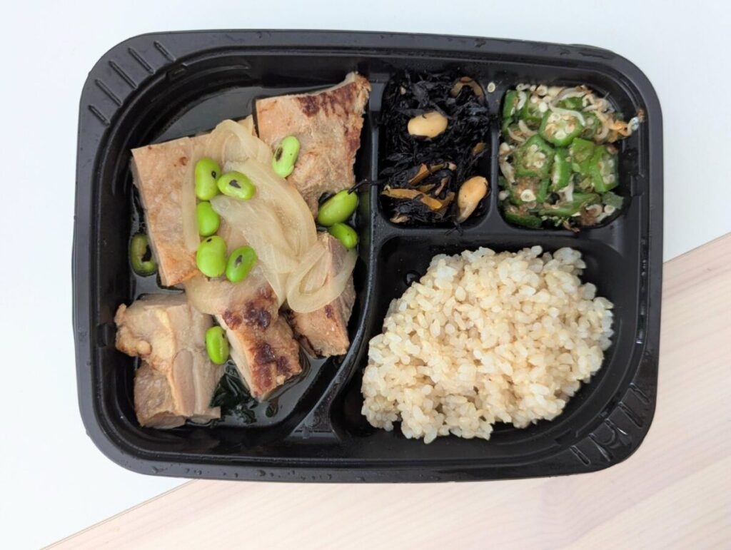 筋肉食堂DELIの冷凍弁当「てりてりに焼いた鶏もも肉の生姜焼きと香り引き立つ炙り枝豆」。ジューシーな鶏もも肉の生姜焼きをメインに、枝豆、玉ねぎ、副菜としてひじき煮やオクラの和え物、玄米ごはんがセットになっている。