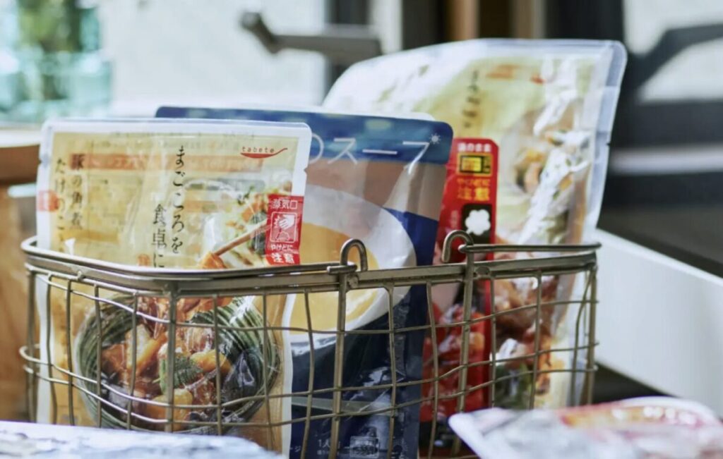 OMA MESIのパウチがワイヤーバスケットに入って並んでいる様子。手前には肉と野菜の煮物、奥にはスープや惣菜パックが見える。