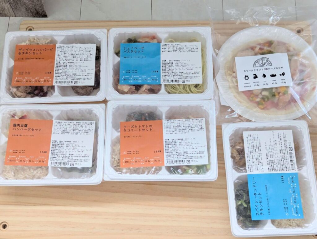 マッスルデリのLEANプラン5食セットとサービスのピザ。 デミグラスハンバーグ＆チキン、ジェノベーゼパスタ、鶏肉豆腐ハンバーグ、チーズとトマトのタコミート、スパイシータンドリーチキン、スモークチキンと彩り野菜チーズピザの6種類が並ぶ。高たんぱく・低脂質なボディメイク向け冷凍弁当。