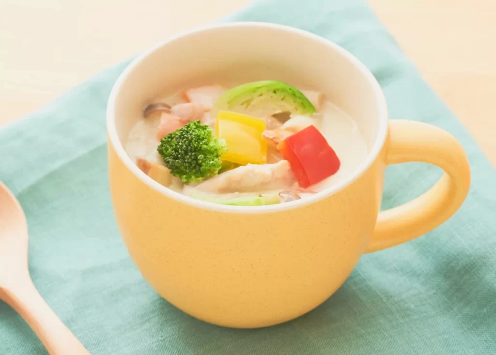 鶏肉・ブロッコリー・パプリカなどが入った、豆乳仕立ての優しい味わいの具だくさんスープ。黄色いマグカップに盛り付けられている様子の写真。