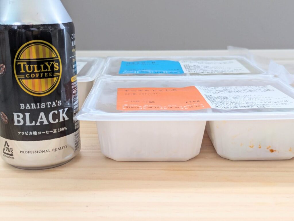 マッスルデリの冷凍弁当容器をTULLY'Sの缶コーヒーと並べて撮影した写真。 容器の高さが缶の約半分程度で、冷凍庫にも収納しやすい厚み。 手前にはオレンジラベル（LEANプラン）の弁当が並ぶ。