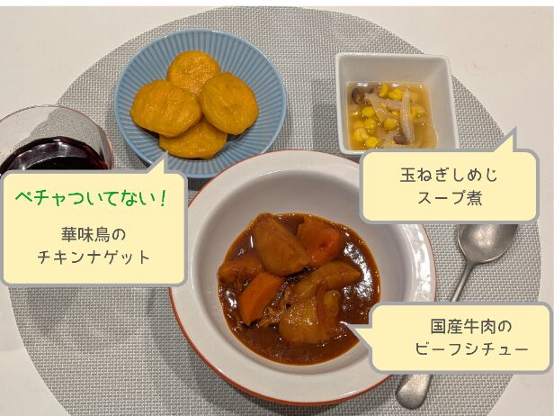 わんまいる『国産牛肉のビーフシチュー』の盛り付け例。中央にビーフシチュー、左に華味鶏のチキンナゲット、右にコーン・玉ねぎ・しめじのスープ煮。優しい色合いで家庭料理風にまとまったセット。