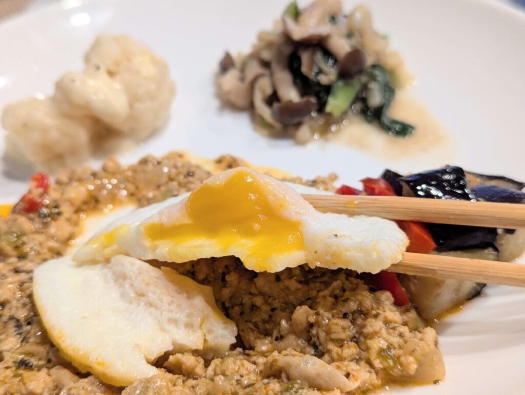 三ツ星ファームガパオの目玉焼きを箸で持ち上げた様子のアップ写真。後ろにはカリフラワーときのこの副菜。
