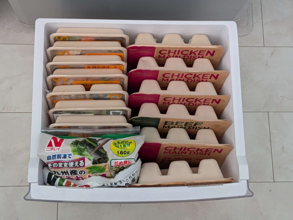 冷凍庫の引き出しに、デリピックスとナッシュ（nosh）の冷凍ミールが整然と並べられている。手前には冷凍ほうれん草のパッケージがあり、奥には『CHICKEN MAIN DISH』や『BEEF MAIN DISH』などと書かれた茶色やピンク色の紙製容器が複数並んでいる。